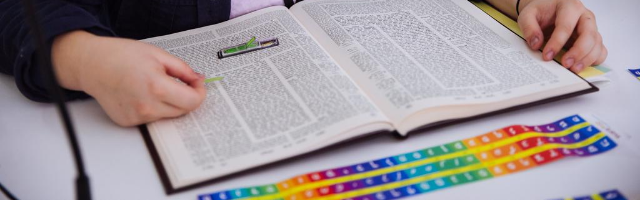 someone is holding an open book of Talmud, with a rainbow strip of the alef-bet laid down on top of the book.